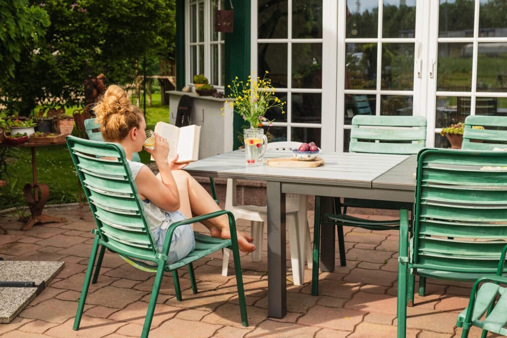 woman drinking from a cup sitting at a table in a lush backyard while reading a book