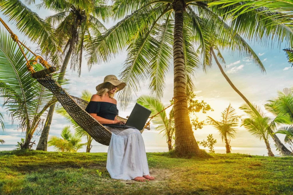 Woman in hat sitting in hammock on the beach with laptop.